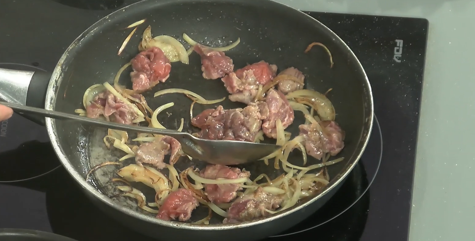 Una sarten oscura con cebolla y carne cruda, junto a una cuchara metalica gris.